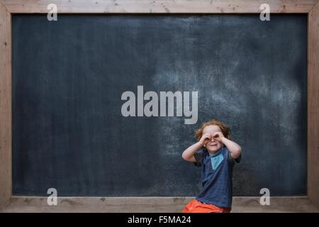 Young boy standing in front of blackboard, faisant de verres avec les mains Banque D'Images