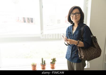 Mature Woman standing in front of window holding smartphone smiling at camera Banque D'Images