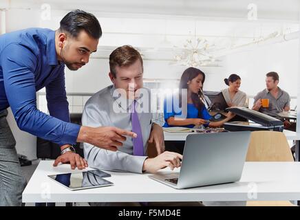 Deux hommes d'avoir un débat, looking at laptop, des collègues qui travaillent en arrière-plan Banque D'Images