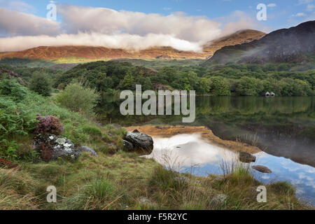 La rive du Llyn Dinas, Snowdonia, est à l'ombre alors que le lever tôt le matin s'allume la nuée couvrit la montagne. Banque D'Images