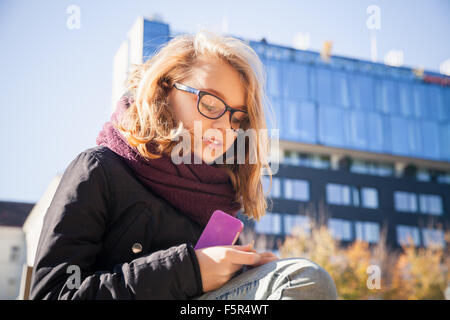 Belle blonde caucasienne adolescente dans les verres assis dans city park et est titulaire d'smartphone dans les mains Banque D'Images