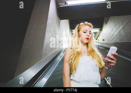 Demi-longueur de jeunes beau young blonde woman on a les cheveux raides escaliers mécaniques dans le métro, la tenue d'un smartphone, à la recherche et à la baisse sur l'écran - la technologie, communication concept Banque D'Images