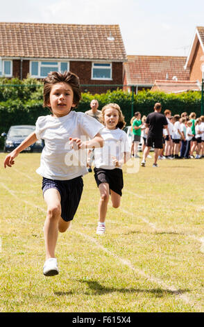 Caucasien enfant, garçon, 8-9 ans. La journée du sport scolaire. Running on grass track vers viewer avec expression excité, avec fille courir derrière dans la course. Banque D'Images