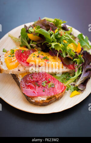 Sandwich ouverts avec heirloom tomates et fromage de chèvre avec un côté de jardin verts, sur un tableau noir haut-fond. Banque D'Images