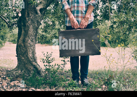 Un jeune homme de race blanche portant une chemise à carreaux porte une vieille valise marron rustique dans un décor naturel, avec un effet de filtre Banque D'Images