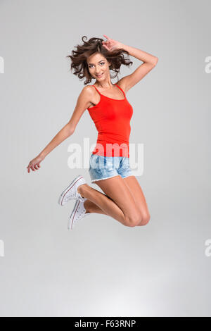 Jeune femme insouciante heureux bouclés à Red top, short jeans blanc et sneackers jumping and smiling Banque D'Images