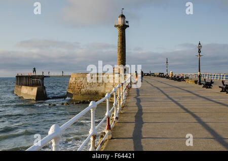 Vue pittoresque de personnes marchant par la mer sur la jetée, Whitby, North Yorkshire (GB) - Soir d'été soleil projette de longues lignes d'ombre d'un phare. Banque D'Images