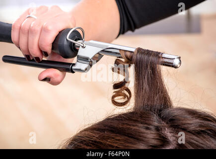 Hairstylist à l'aide d'un fer à friser ou des pinces à ferme anglaises dans les longs cheveux bruns d'une cliente Banque D'Images