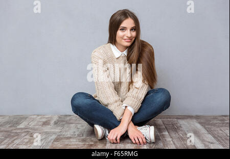 Beautiful smiling girl with long hair en beige pull et jeans assis sur un plancher en bois avec les jambes croisées Banque D'Images