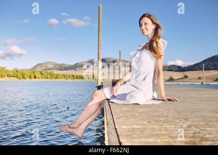 Portrait d'une femme blonde assise sur un quai sur un lac, avec une robe blanche Banque D'Images