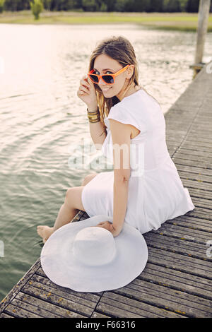 Portrait d'une femme assis sur un quai avec robe blanche et chapeau et verre orange Banque D'Images