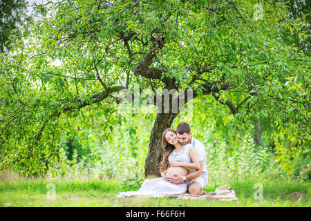 Happy young couple expecting baby, femme enceinte avec mari toucher ventre, assis sur l'herbe verte Banque D'Images