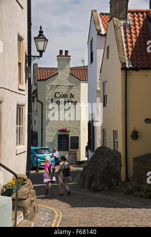 Les visiteurs marchent vers le bas le pittoresque, étroites et pavées, High Street, dans le village traditionnel de pêcheurs de Staithes, North Yorkshire, UK, sur une journée ensoleillée. Banque D'Images