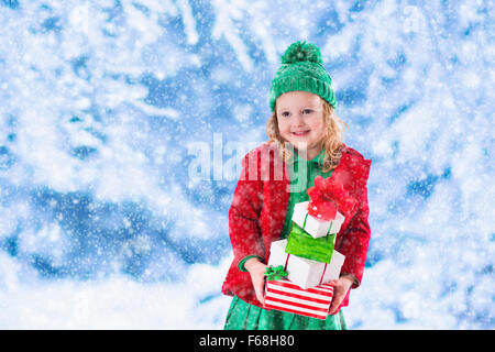 Petite fille en rouge et vert étoffes hat holding Christmas present cases dans Winter Park sur la veille de Noël. Les enfants jouent dans la neige en plein air Banque D'Images