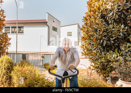 Personnes âgées de sexe masculin ne cesse de monter par l'octogénaire doing exercise bike sur le jardin de la maison Banque D'Images