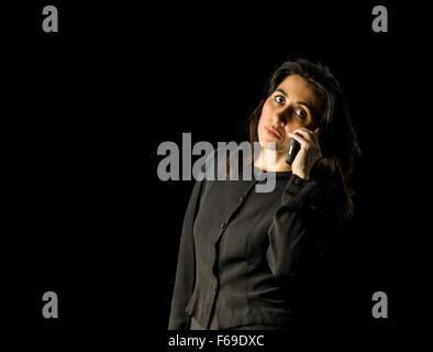 Femme brune en robe noire, debout devant un fond noir et la tenue d'un téléphone cellulaire jusqu'à son oreille. Banque D'Images