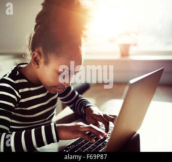 Jeune fille noire avec un chignon afro concentrant comme elle tape sur un ordinateur portable, Close up side view avec sun flare retour Banque D'Images