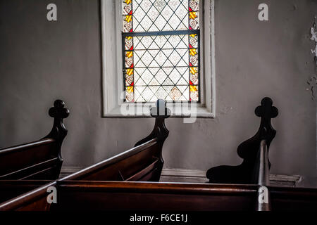 Une pièce faiblement éclairée de l'intérieur de l'église un vitrail fenêtre avec croix sculptées décorativement mis sur les extrémités de chaque art pew en bois Banque D'Images