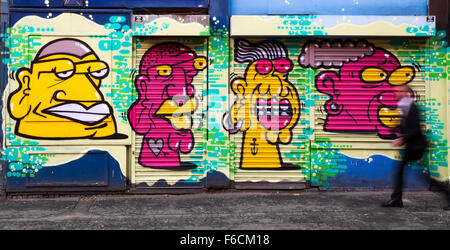 Portraits de visages de dessins animés mauves jaunes ; femme passant des volets roulants décorés, couleurs vives représentant divers visages de personnages de dessins animés. Manchester, Royaume-Uni Banque D'Images