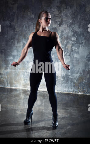 Forte femme bodybuilder dans les vêtements noirs sur fond de mur. Banque D'Images