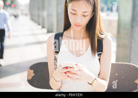 La moitié de la longueur de cheveux longs asiatique beau jeune patineuse femme marche dans la ville, à l'écoute de la musique avec des écouteurs, holding smartphone à la recherche vers le bas et en tapotant sur l'écran - la musique, technologie concept Banque D'Images