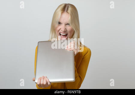 Jeune femme blonde au visage en colère montrant l'appareil photo tout en maintenant son ordinateur portable contre l'arrière-plan blanc. Banque D'Images