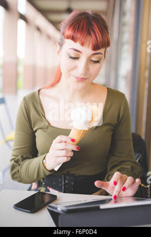 Beau portrait jeune femme rousse assis dans un bar à l'aide de tablet en mangeant de la crème glacée, à la technologie, à la baisse - réseau social, concept multitâche - portant chemise verte et floral jupe Banque D'Images