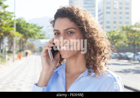 Belle femme latine avec chemisier bleu au téléphone dans une ville d'Amérique latine avec des bâtiments modernes et arbres en arrière-plan Banque D'Images