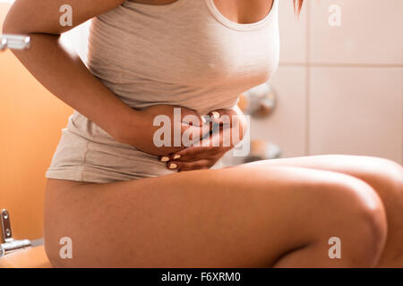 Close up malade Jeune femme assise sur les toilettes dans la salle de bains tout en tenant son estomac douloureux Banque D'Images