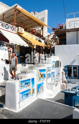 Santorin, Fira. Art shop souvenirs avec affichage des peintures de Santorin sur mur, avec les touristes en marche. Arrière-plan, taverne sur terrasse. Banque D'Images