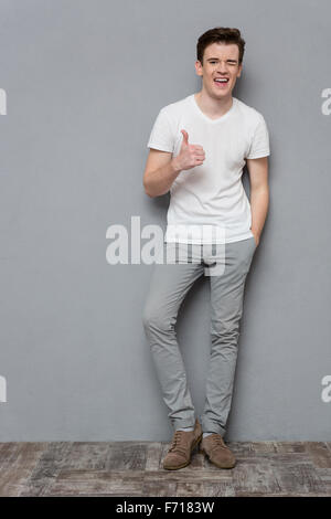 Full Length portrait of smiling mature inspiré certain jeune homme en t-shirt blanc et un pantalon gris montrant jusqu'thimbs et gagnez Banque D'Images