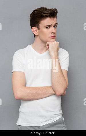 Beau jeune homme pensif attrayant en blanc T-shirt looking at camera, tenant la main sur le menton et le dos gris contre permanent Banque D'Images