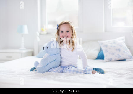 Funny happy girl reading a book et jouer avec son jouet ours en peluche dans le lit. Les enfants jouent à la maison. Pépinière blanc. Banque D'Images
