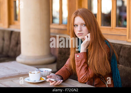 Belle jeune femme rousse aux cheveux longs en blouson de cuir à l'aide de téléphone cellulaire et d'attendre que quelqu'un dans le café en plein air Banque D'Images