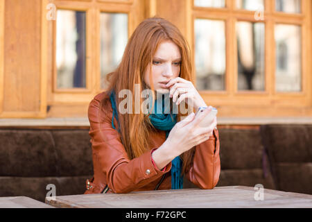 Embarrassé triste shoked jeune rousse dame lit un message sur téléphone mobile assis dans un café en plein air Banque D'Images