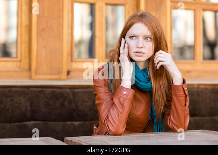 Inquiets concerné jeune rousse femme en blouson de cuir et foulard talking on cellphone in outdoor cafe Banque D'Images