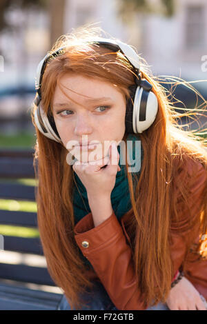 Portrait de jeune femme rêveuse triste magnifique avec de longs cheveux rouges dans les écouteurs assis dans le parc Banque D'Images
