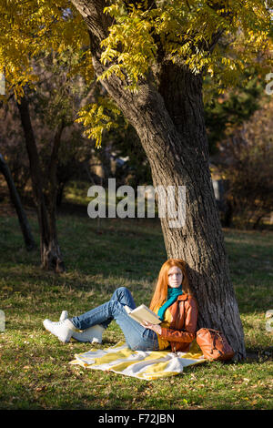 Young redhead femme lisant un livre assis sous l'arbre dans le parc et de rêver Banque D'Images