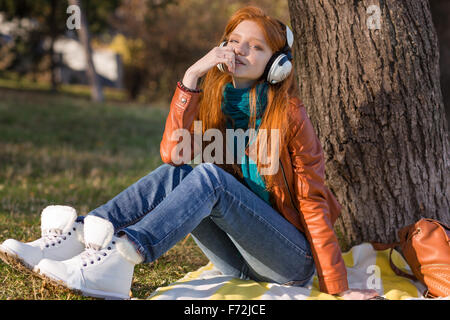 Jolie jeune femme pensive en veste de cuir, des bottes blanches et jeans écouter de la musique sous l'arbre Banque D'Images