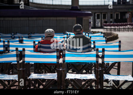 Couple assis dans des chaises à rayures bleues et blanches à l'extérieur dans le soleil à Eastbourne bandstand, Eastbourne, East Sussex, England, UK Banque D'Images