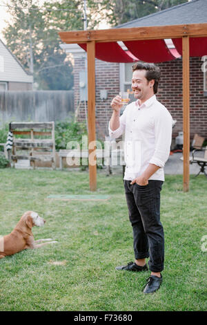 Homme debout dans un jardin, prendre un verre, un chien couché dans l'herbe à côté de lui. Banque D'Images