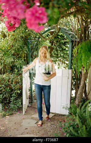 Blonde qui entre dans un jardin par une porte en bois blanc, porteur d'une plante en pot. Banque D'Images