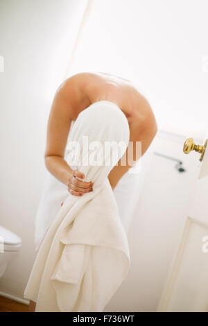 Femme enveloppée dans une serviette blanche debout dans une salle de bains, enveloppant ses cheveux dans une serviette. Banque D'Images