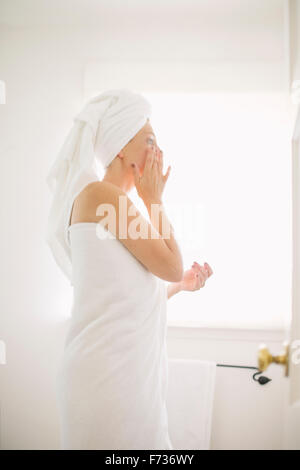 Femme enveloppée dans une serviette blanche debout dans une salle de bain, l'application de la crème. Banque D'Images