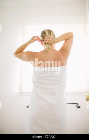 Femme enveloppée dans une serviette blanche debout dans une salle de bains, faire ses cheveux mouillés. Banque D'Images