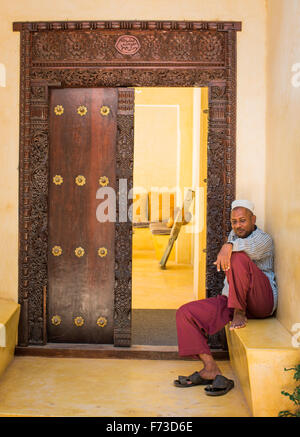 LAMU, Kenya, Afrique. Un homme en robe arabe traditionnelle se trouve sur un becnh à l'extérieur une ancienne porte en bois sculpté. Banque D'Images