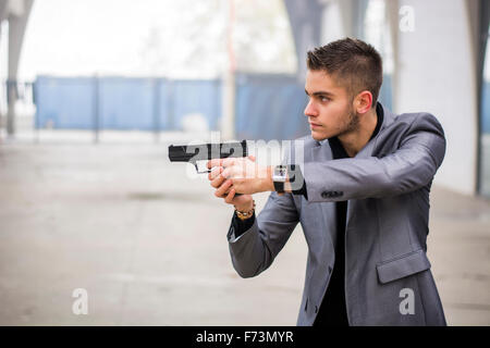 Bien habillé beau jeune détective ou policier ou gangster debout dans un environnement urbain visant une arme à feu sur la gauche Banque D'Images