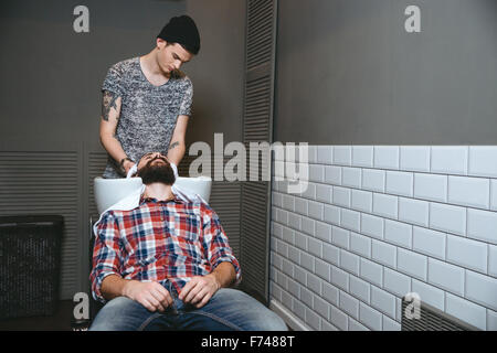 Jeune beau salon de coiffure avec chapeau noir tatoo dans le lavage des cheveux du client avec barbe en chemise à carreaux et jeans dans barbershop Banque D'Images