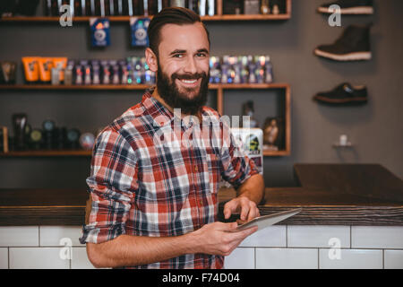 Bel homme heureux avec barbe en chemise à carreaux à l'aide de tablet dans barbershop Banque D'Images