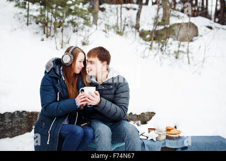 Heureux Couple Sitting Outdoors in Winter Forest de boire une boisson chaude et à la recherche à l'autre Banque D'Images